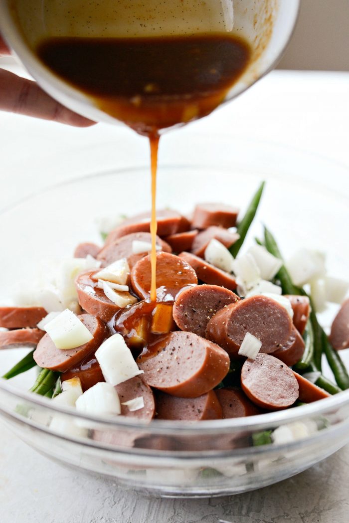 bowl of sausage, onion, green beans, salt and pepper with barbecue sauce being poured on top.