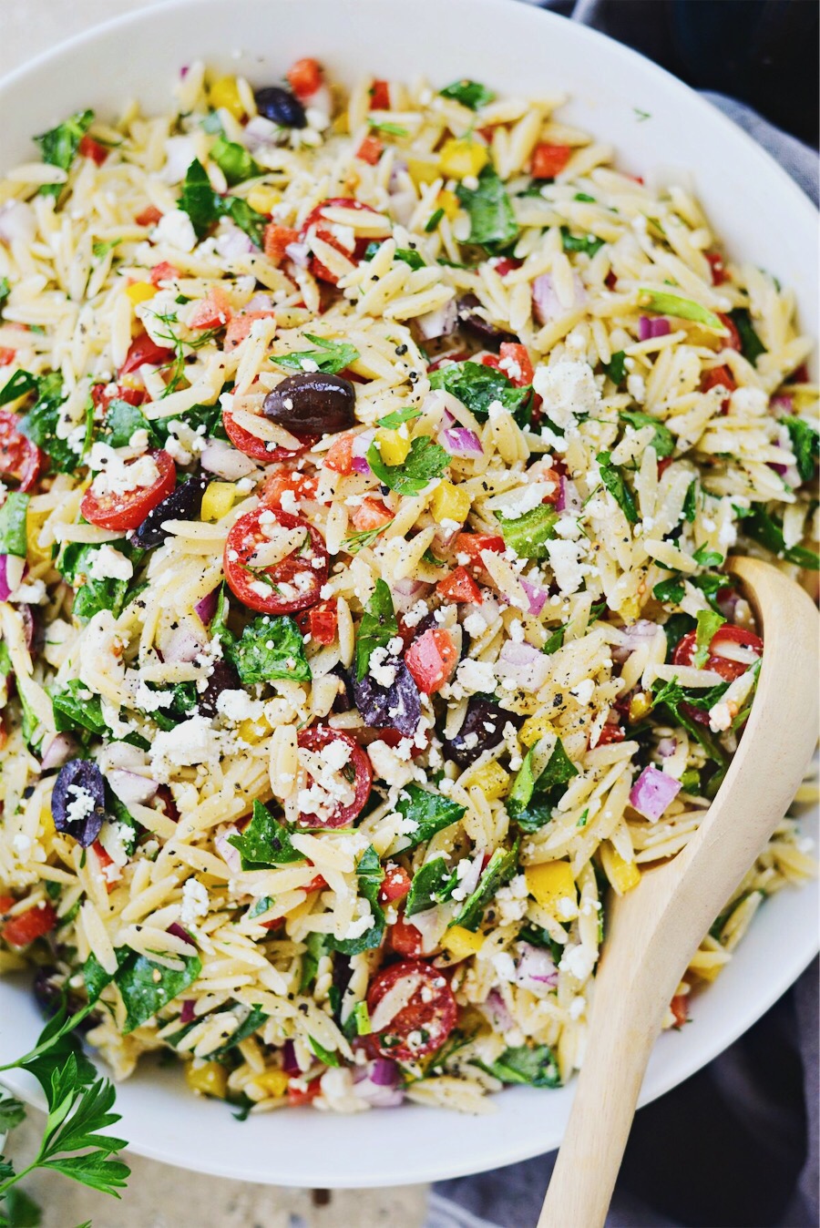 Greek Orzo Salad served in white bowl with wooden spoon.