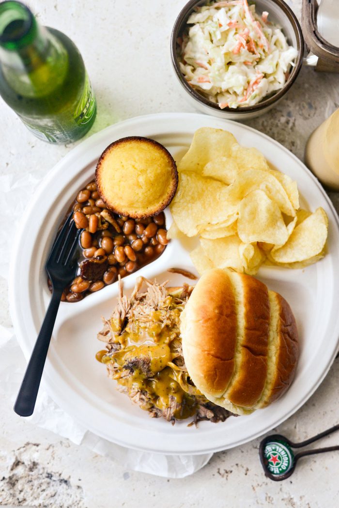 Plate of pulled smoked pork on brioche bun with beans, chips, cornbread and coleslaw.